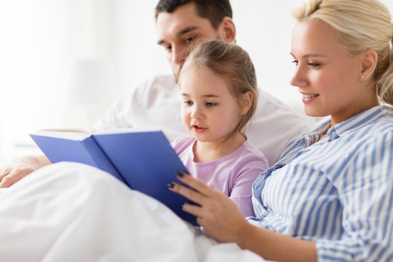 Mom, dad, and daughter read a blue book under white covers in their Beaufort, NC, home.