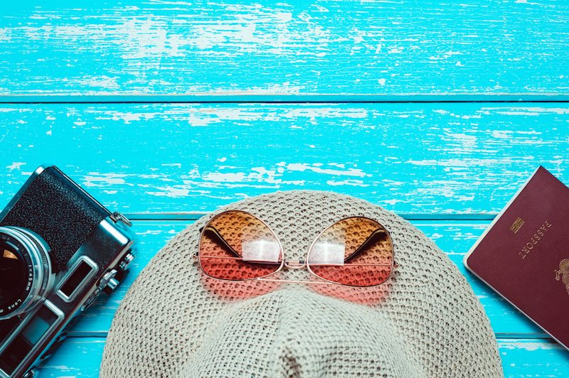 Camera, sun hat, sunglasses, and a passport on a blue picnic table in a summer home, where the AC is blowing hot air.