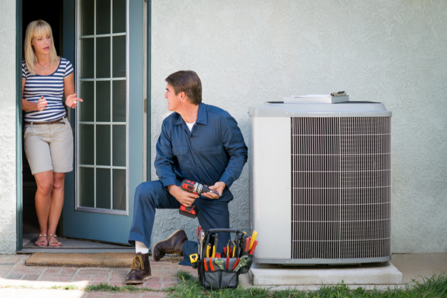 This picture represents a man who is fixing up an air conditioner while talking to the homeowner.