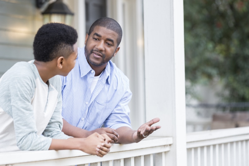 How Does a Gas Furnace Work? A father talks with his hands as he leans against the railing of his front porch with his preteen son and has a serious discussion.