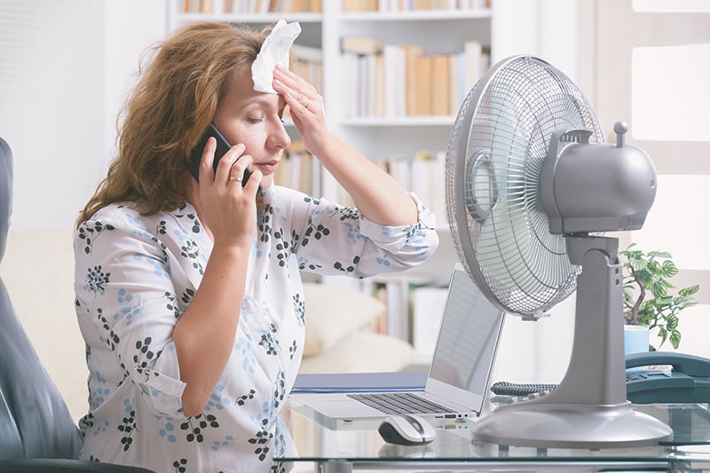 When Do I Replace My Air Conditioner? A woman sitting in front of a fan trying to cool off.