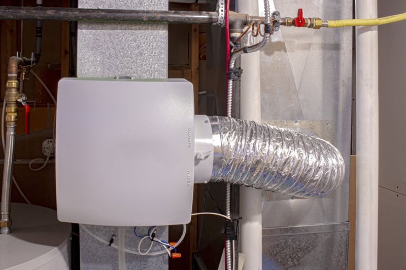 A white rectangular bypass humidifier mounted onto galvanized metal ductwork, connected to a flexible foil duct that loops to an adjacent duct section.